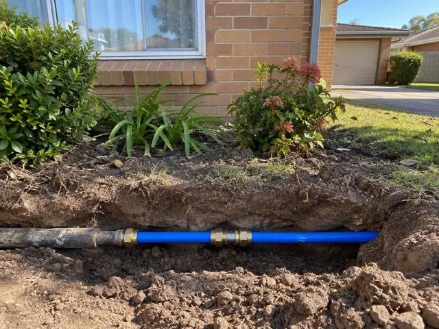 Exposed repaired water pipe in a garden trench beside a brick house in Brookvale